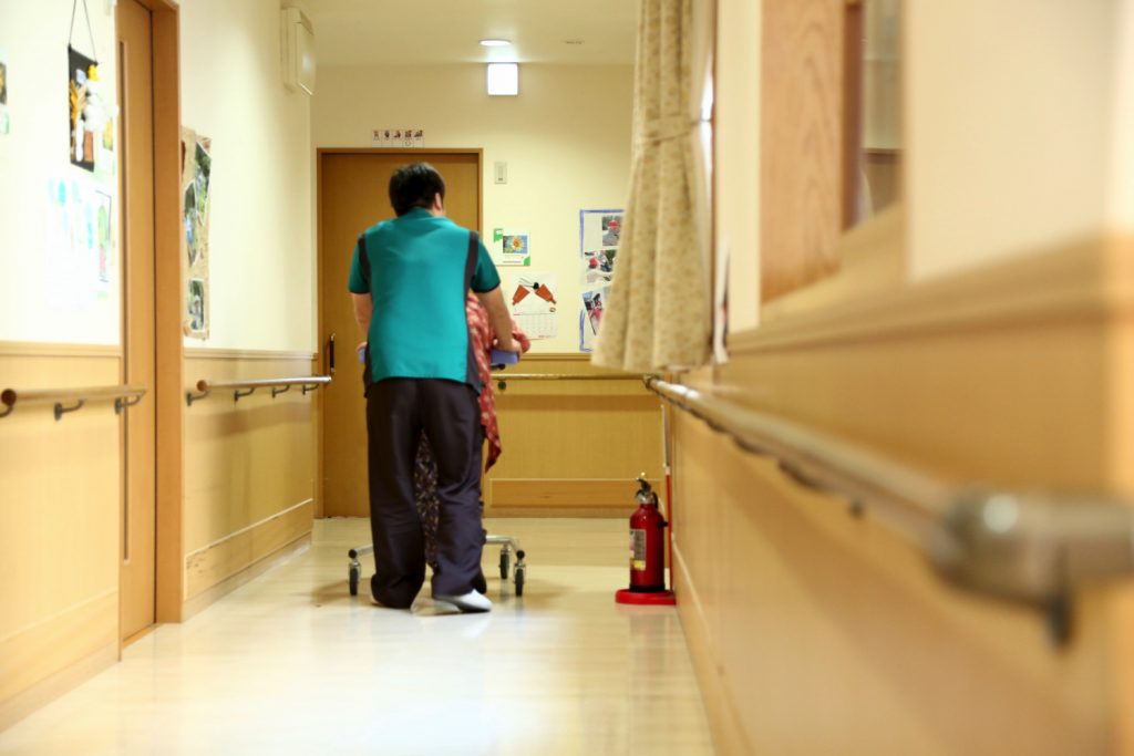 廊下を歩く入居者と介護スタッフ、掲示物や手すりが整備された通路